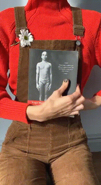 Человек в красном свитере и коричневом комбинезоне держит книгу под названием «Йога Мала», украшенную белым цветком на бретельке.