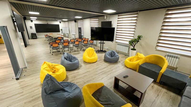 A spacious, modern room featuring distinct zones: a formal area with a long table, chairs, and a large screen, and an informal lounge area with yellow and gray bean bags, modular seating, a coffee table, and a large TV.