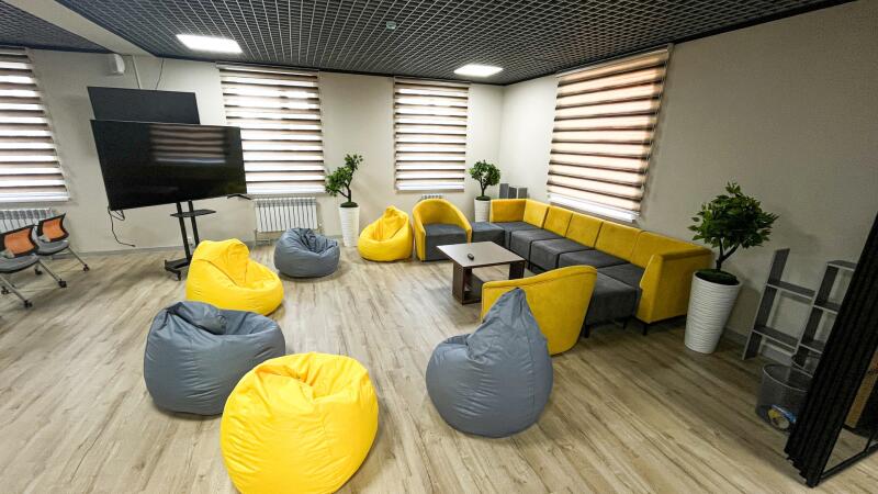 A modern room with light wooden flooring features a yellow and grey modular sofa, yellow armchairs, and numerous yellow and grey beanbag chairs arranged around a brown coffee table. A large black TV screen on a stand is visible on the left, with windows covered by horizontal blinds on the walls.