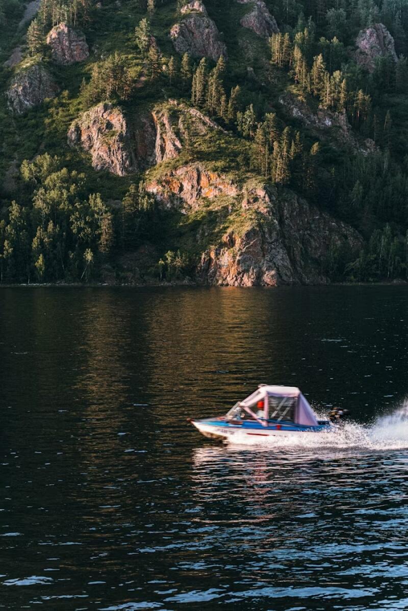 A motorboat speeds across the water in front of a rocky, tree-covered hillside.