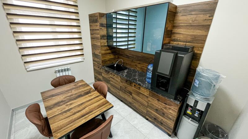 A modern room features a wooden table with brown chairs, a wooden cabinet unit with a black marble-patterned countertop, a sink, and a large mirror. A coffee machine, a water dispenser, and a window with horizontal blinds are also visible.