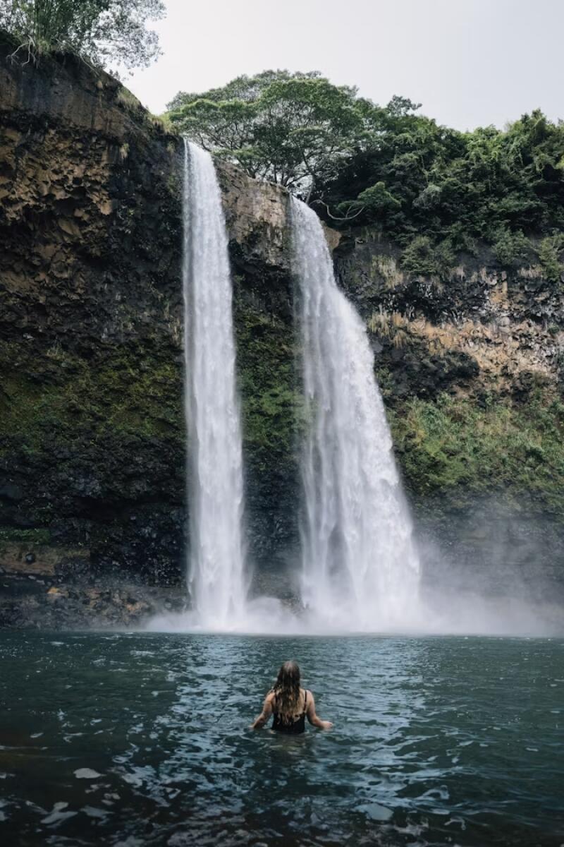 A woman stands in water in front of a tall, rocky cliff with twin waterfalls cascading into the pool, surrounded by lush green vegetation.