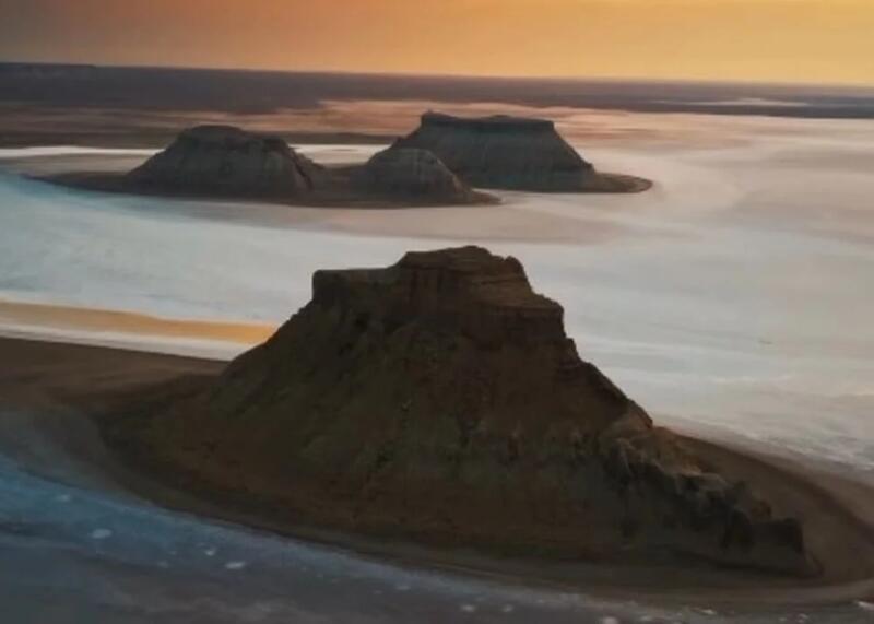An aerial view of a desert landscape featuring three dark, flat-topped hills rising from a light-colored, possibly salt-covered plain under an orange and blue sky.