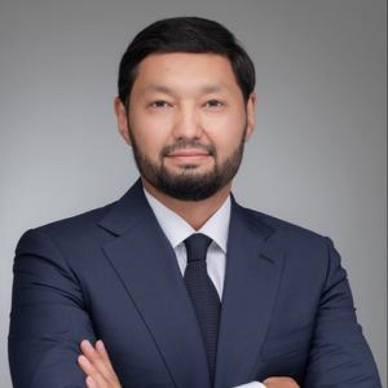 A man with a beard in a navy suit and tie is posing with his arms crossed against a gray background.
