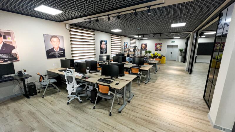 A modern computer lab or office space with rows of desks, desktop computers, and chairs. Light-colored walls display posters of people, windows have blinds, and a grid ceiling with lights illuminates the wooden floor. A reflection on a glass partition shows 'Sardahu Student Center'.