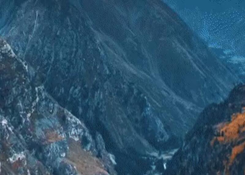 A high-angle view of a deep mountain valley surrounded by steep, dark blue-grey peaks, with patches of green and brown vegetation visible on the slopes.