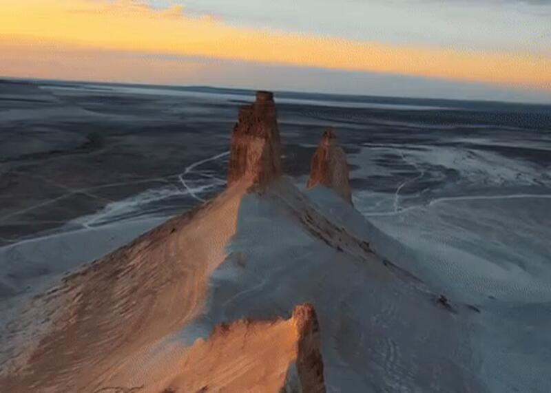 An aerial view of a desert landscape featuring several prominent rock formations with warm light on their peaks, set against a vast flat plain with tracks and a sunset sky.