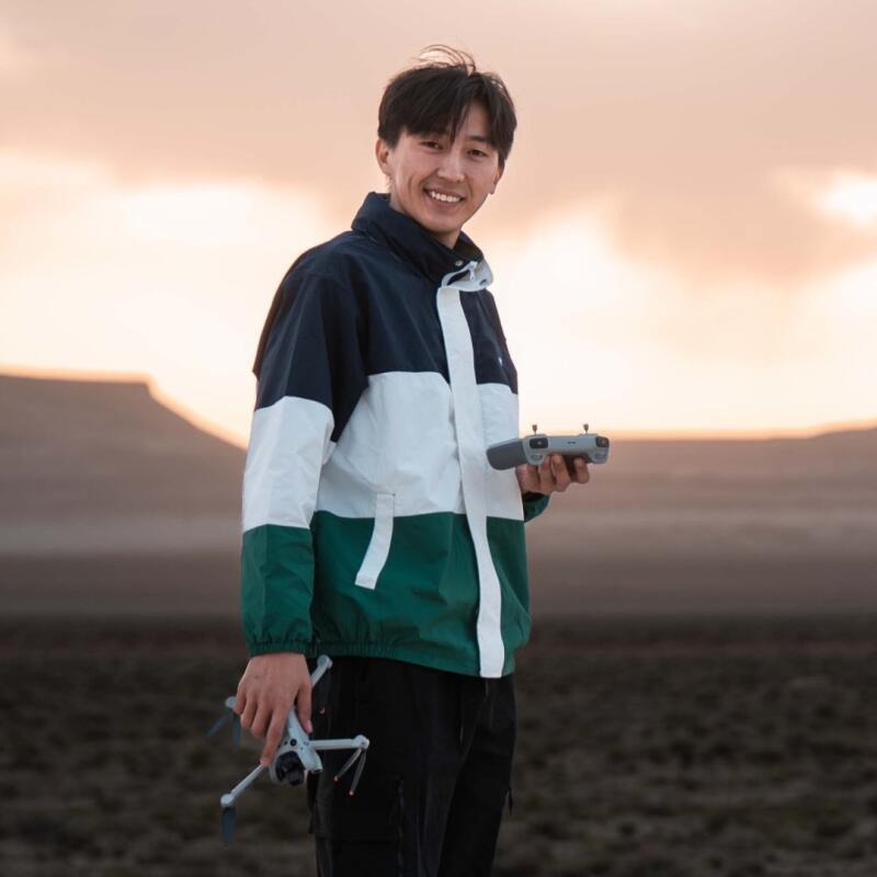 A smiling person in a tri-color jacket stands outdoors, holding a white drone in their left hand and its grey remote control in their right hand, with a warm-toned sky and distant hills in the background.