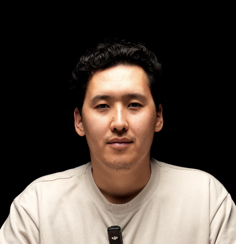 A man with dark, curly hair and a light beige shirt looks directly at the camera against a black background, with a black DJI microphone clipped to his shirt.