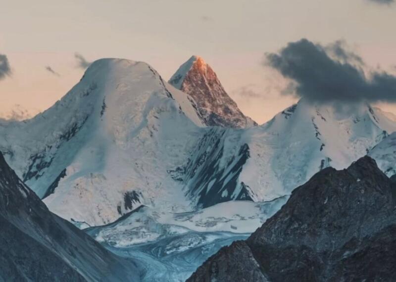 Snow-covered mountains and glaciers are visible under a pale sky, with one central peak illuminated by golden light and dark clouds on the right.