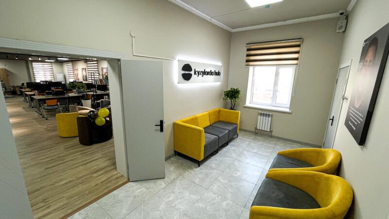 A modern office or co-working space with a waiting area featuring yellow and gray seating, a lighted sign on the wall displaying "kyzylorda hub", and a doorway leading to a room with desks and computers.