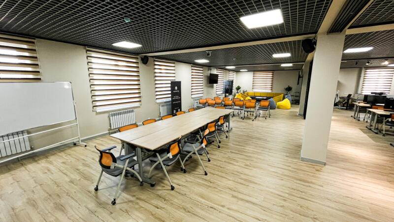 A large, modern multi-purpose room with a long conference table and orange chairs, a presentation area, and a lounge section with a yellow sofa. The room features light wood flooring, a dark grid ceiling, and windows with striped blinds.