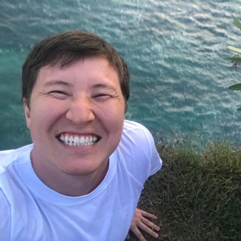 A man in a white t-shirt smiles broadly with closed eyes, showing his teeth, while taking a selfie above blue water and green grass.