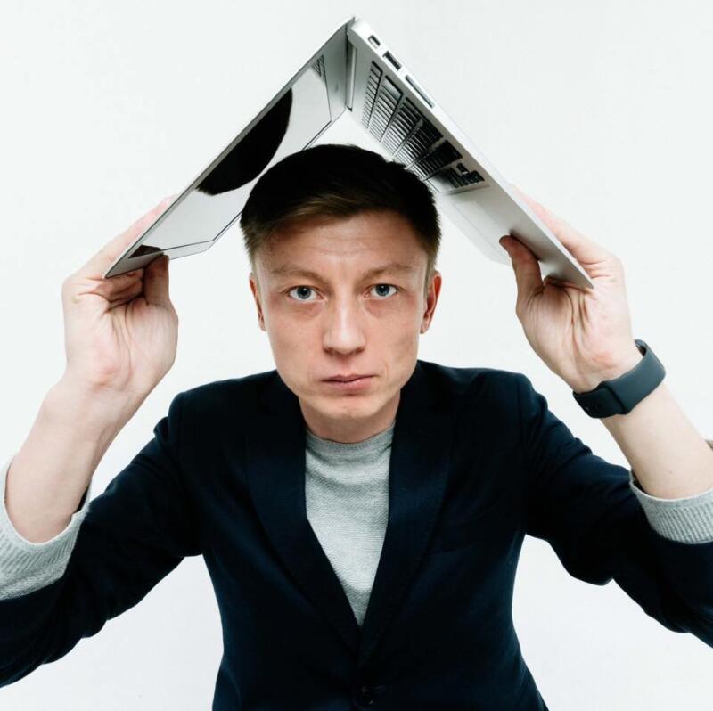 A man in a blazer and gray sweater is holding a silver laptop over his head.