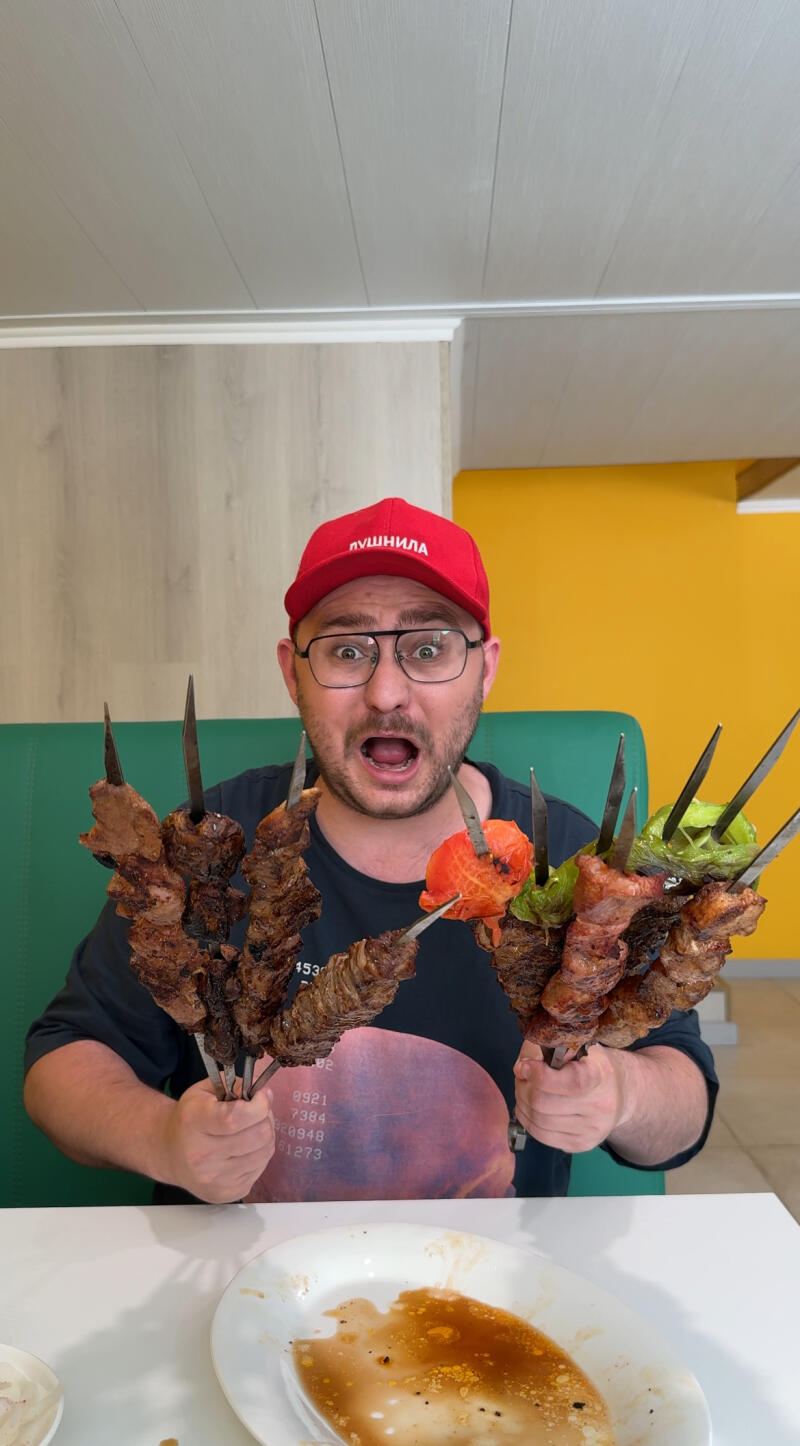 A man with glasses and a red cap with the inscription "Лучший" is holding skewers with grilled meat and vegetables in both hands with an open mouth, standing behind a table with a plate.