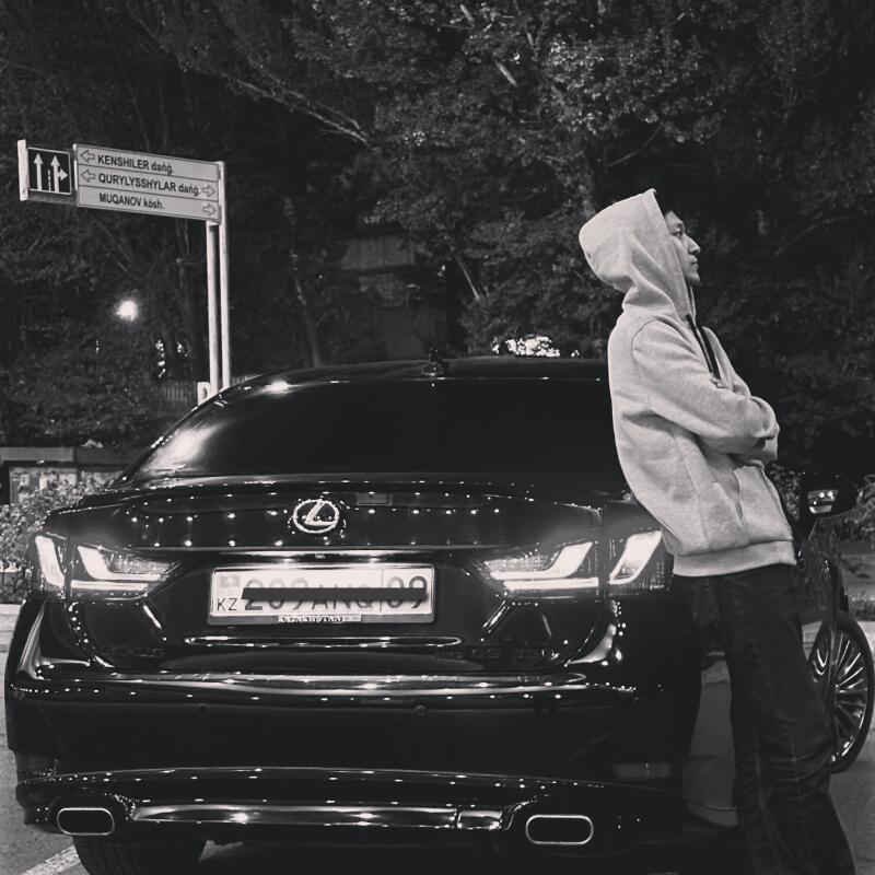 A grayscale shot shows a man in a hoodie leaning against the back of a black Lexus with the license plate "KZ 209 ANG 09", next to a street sign indicating directions to "KENSHILER dańǵ", "QURYLYSSHYLAR dańǵ", and "MUQANOV kósі."