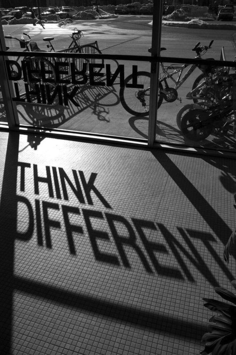 A black and white photo shows the shadow of the words “THINK DIFFERENT” on a tiled floor, cast from a display on a window with bicycles parked outside.