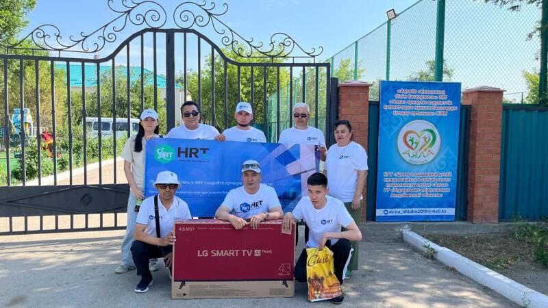 Группа людей стоит на улице с баннером, на котором написано "HRT Создайте лучшее", коробкой от телевизора LG Smart TV и знаком с текстом на казахском и русском языках.