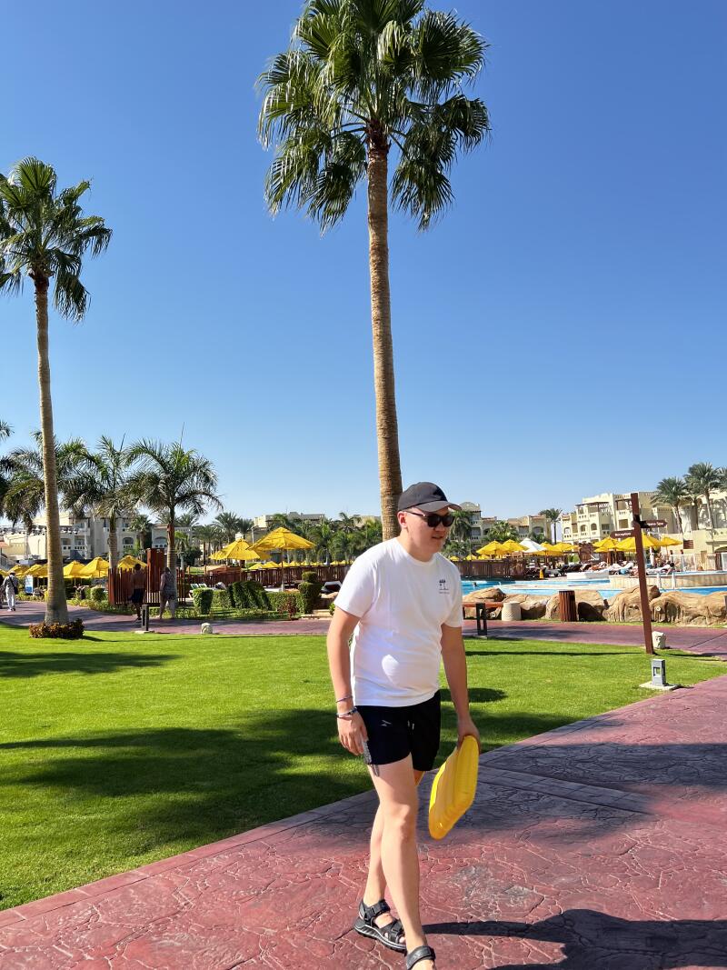 A man in a white t-shirt, black shorts, and sandals walks on a red brick path, carrying a yellow floatation device, with palm trees and a resort in the background under a clear blue sky.