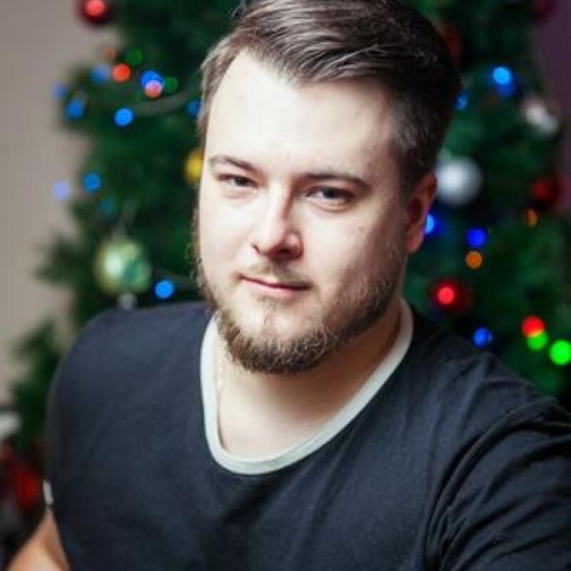 A man with a beard is in front of a Christmas tree with colorful lights.
