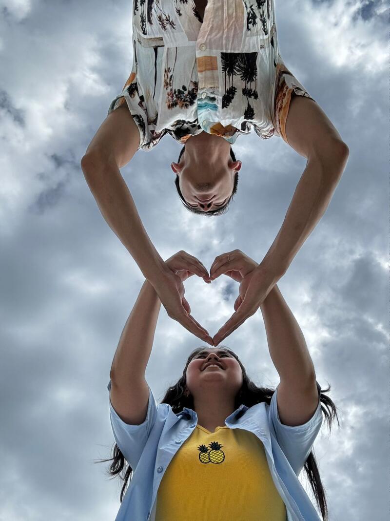 A man and a woman are creating a heart shape with their hands against a cloudy sky, with the man positioned upside down above the woman.