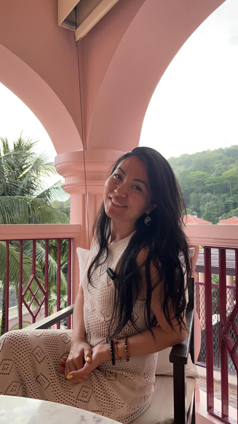 A smiling woman with long dark hair is sitting on a chair on a balcony with pink arches and a view of trees and buildings.