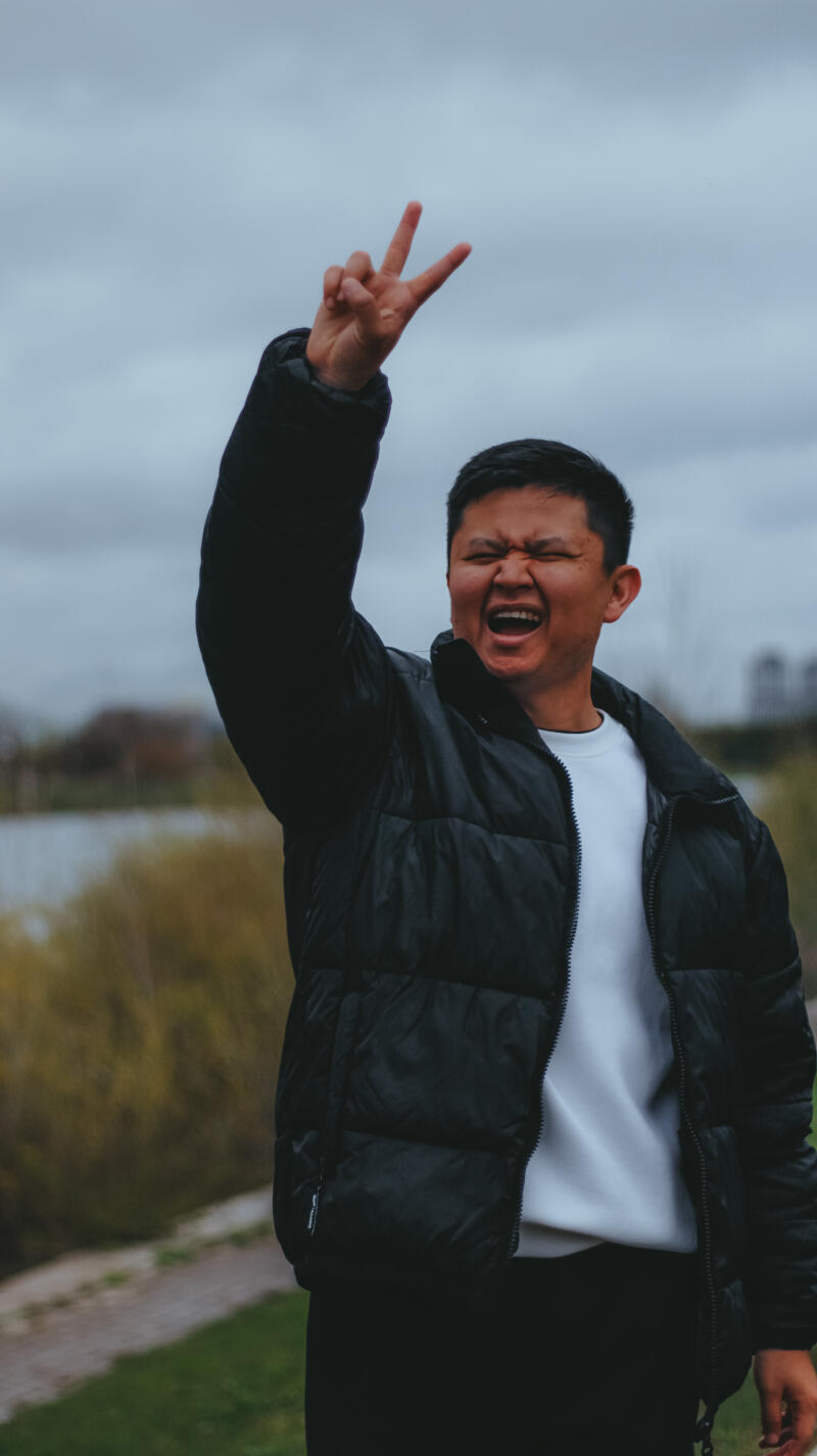 A man wearing a black puffer jacket and a white sweater is smiling and holding up a hand with two fingers raised in a V sign.