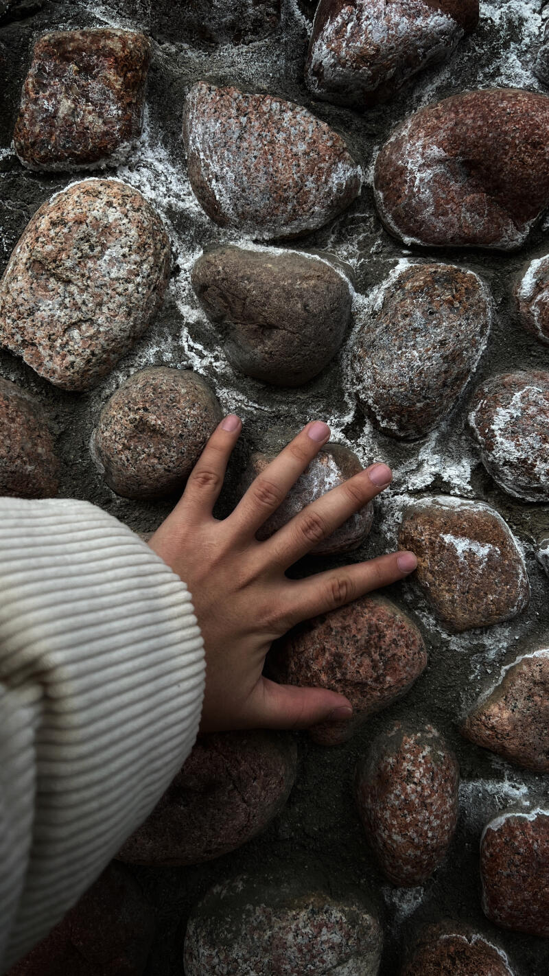 A person's hand touches a wall made of rough, rounded stones, surrounded by gray grout with white residue.