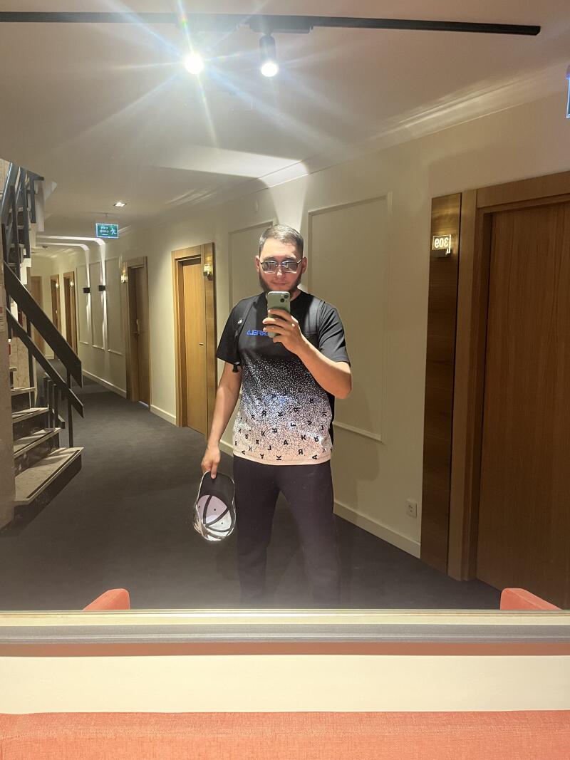 A man with glasses is taking a selfie in a hotel hallway, holding a cap in his hand and standing in front of a mirror.