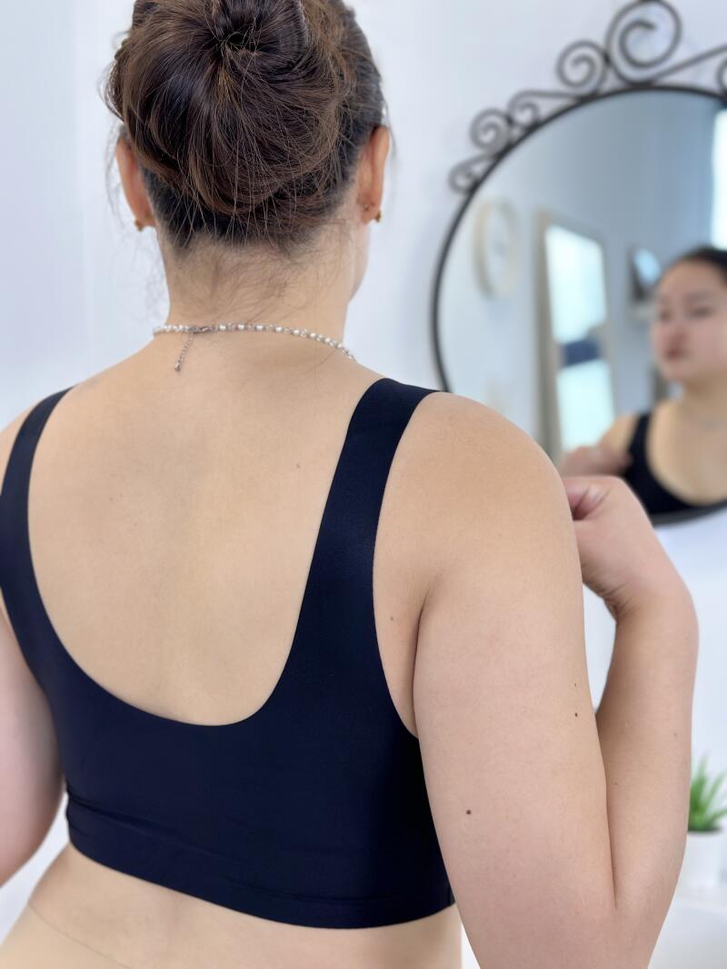 A woman with her hair in a bun is wearing a black sports bra and a pearl necklace while looking in a mirror.