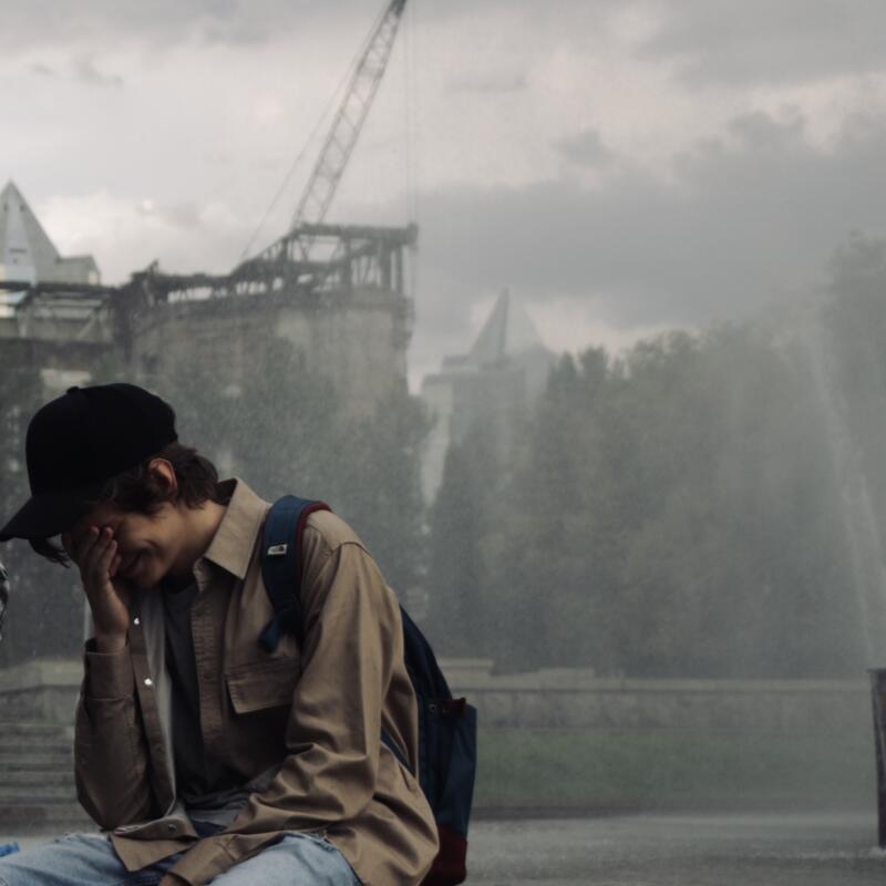 A person in a black cap and a tan shirt sits with their hand on their face near a fountain and a building with a crane in the background.