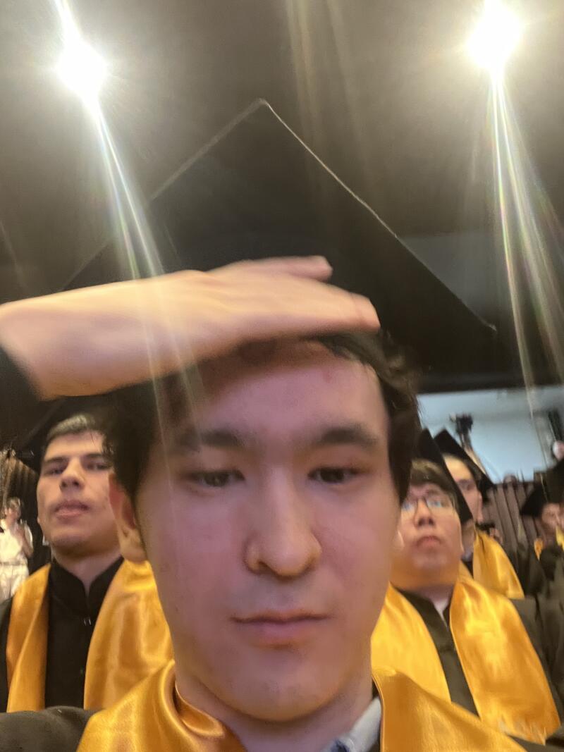 A selfie of a young man in a graduation gown and mortarboard, with other graduates visible in the background.