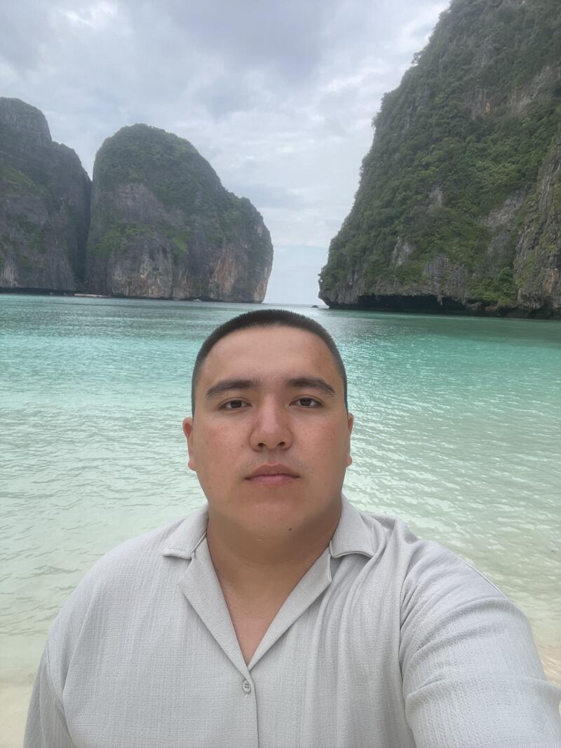 Мужчина в серой рубашке стоит на пляже с бирюзовой водой, на фоне которого видны большие скалистые острова под облачным небом.