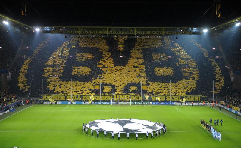 A soccer stadium is filled with fans holding yellow cards to form the BVB logo, with a large soccer ball banner on the field and the text "KEINER SOLLTE ES WAGEN UNS DEN BVB SCHLAGEN!" displayed.