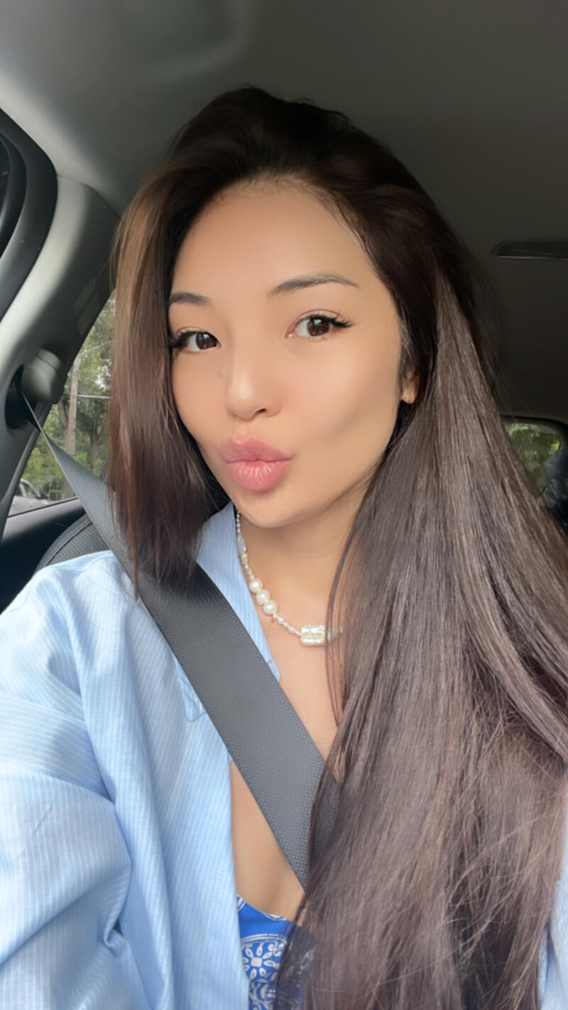 A woman with long brown hair is wearing a blue shirt, a pearl necklace, and a seatbelt.