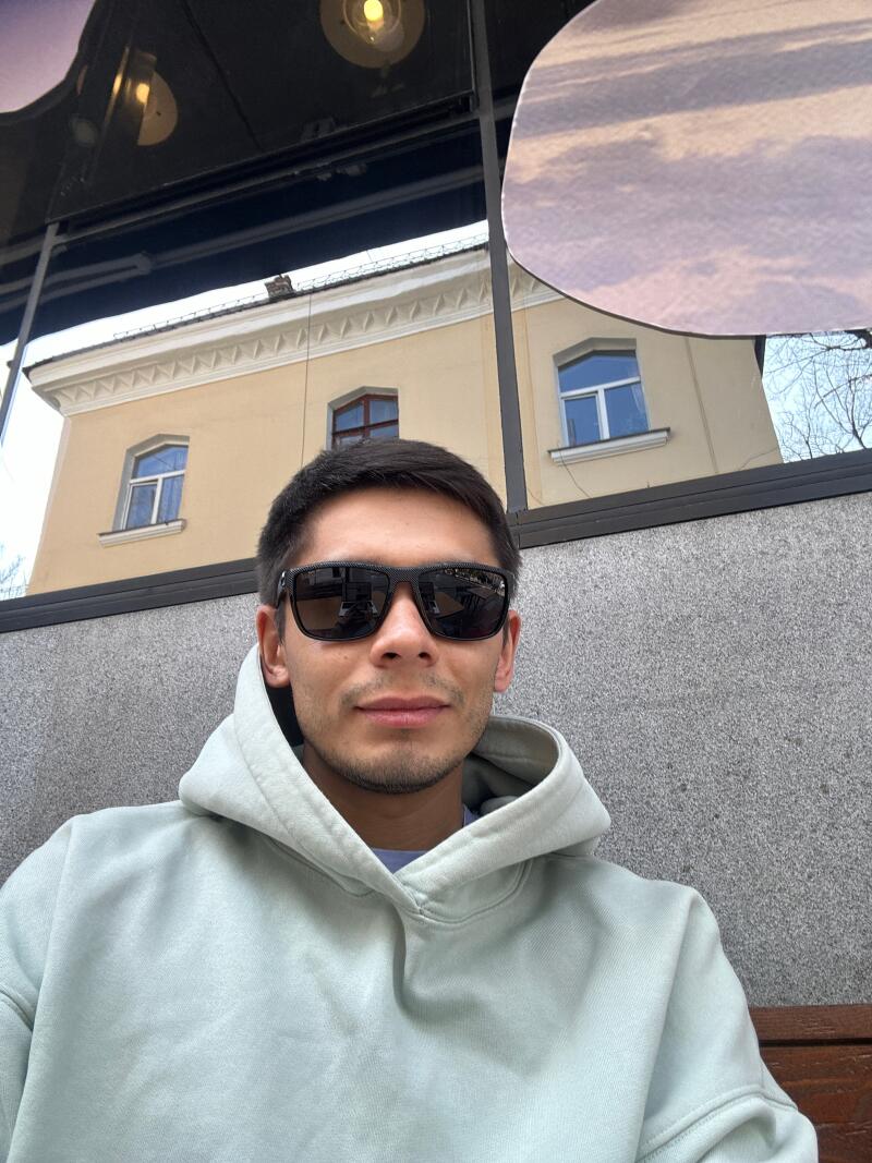 A man with sunglasses and a hoodie is sitting outside with a building reflected in the glass behind him.