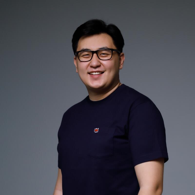 A smiling Asian man with glasses and a navy blue t-shirt stands against a gray background.