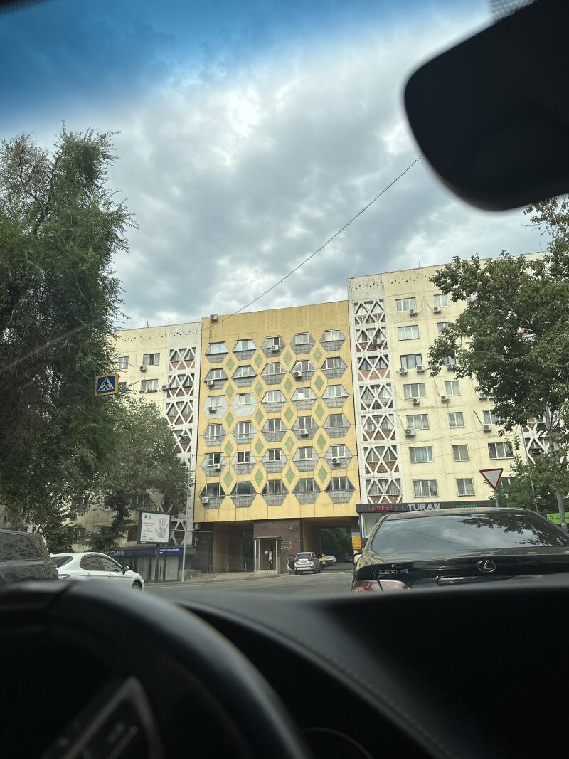 The photo shows a view from inside a car, looking out at a street with buildings, trees, and other vehicles; the building in the center has a distinctive geometric pattern on its facade and a sign that reads "TURAN".