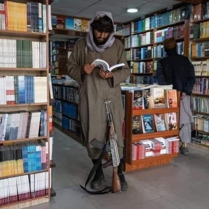 A man in a headscarf and traditional clothing is reading a book in a bookstore, with an assault rifle leaning against his leg.