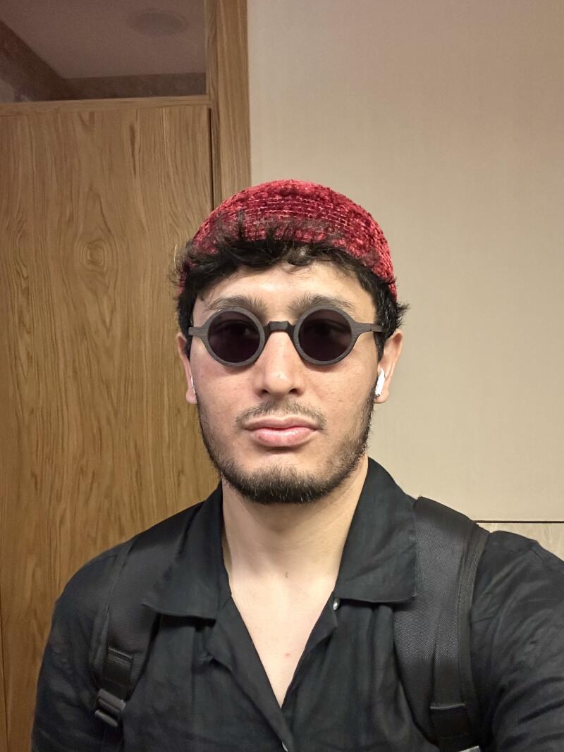 A man with a beard wears a red beanie and round sunglasses, with wireless earbuds in his ears, and a black button-down shirt with a backpack.