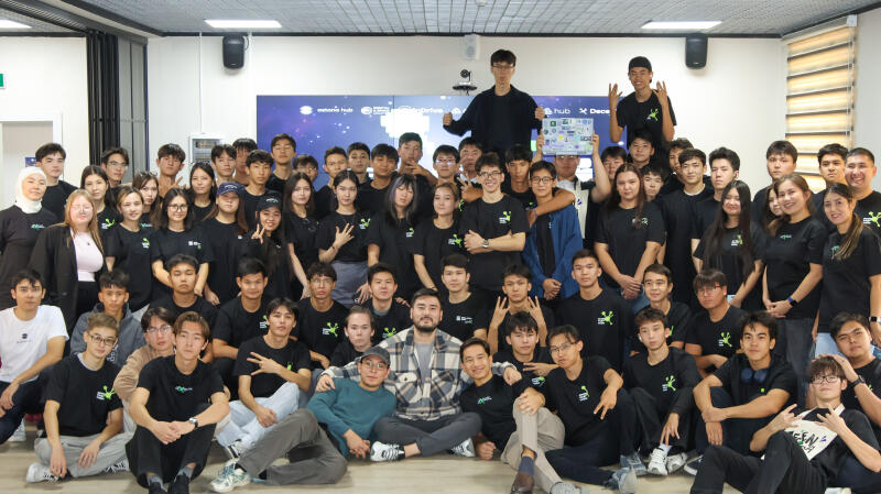 A large group of young people, mostly wearing black T-shirts with a green logo, are posing for a photo indoors, some holding up peace signs, with a screen showing logos including "Astana Hub" in the background.