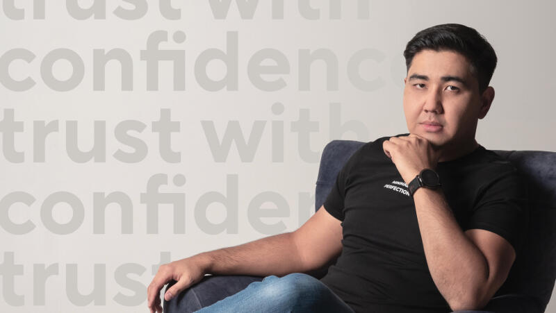 A man in a black t-shirt and jeans sits in a blue chair, with the words "trust with confidence" repeated in the background.