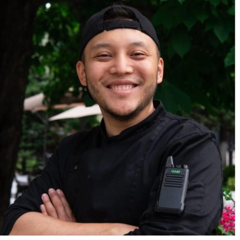 A smiling man in a black chef's jacket and a backward black cap, with crossed arms and a walkie-talkie labeled "WAV" on his sleeve, stands outdoors.