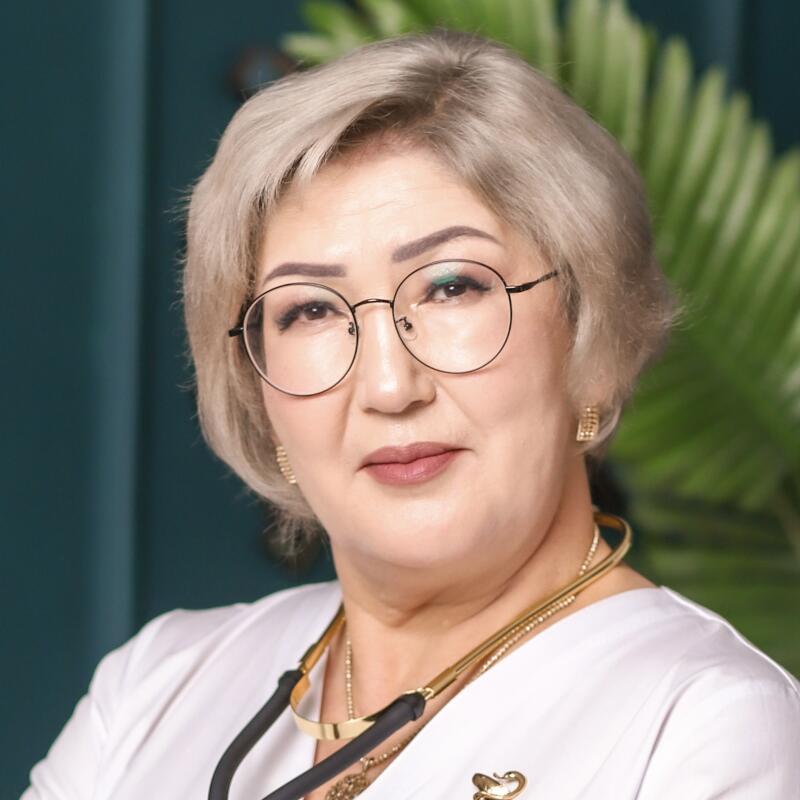 A portrait of a woman with short gray hair and glasses, wearing a white coat with a stethoscope around her neck and gold jewelry.