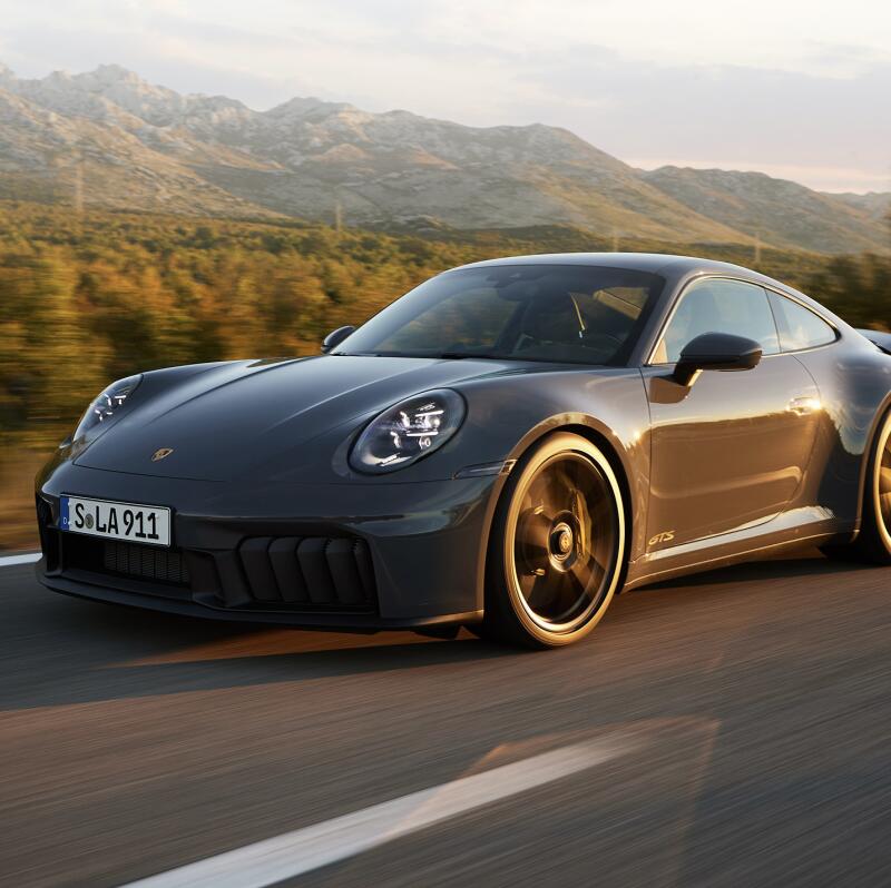 A gray Porsche 911, with license plate 'S.LA 911', is in motion on a paved road, with blurred mountains in the background.