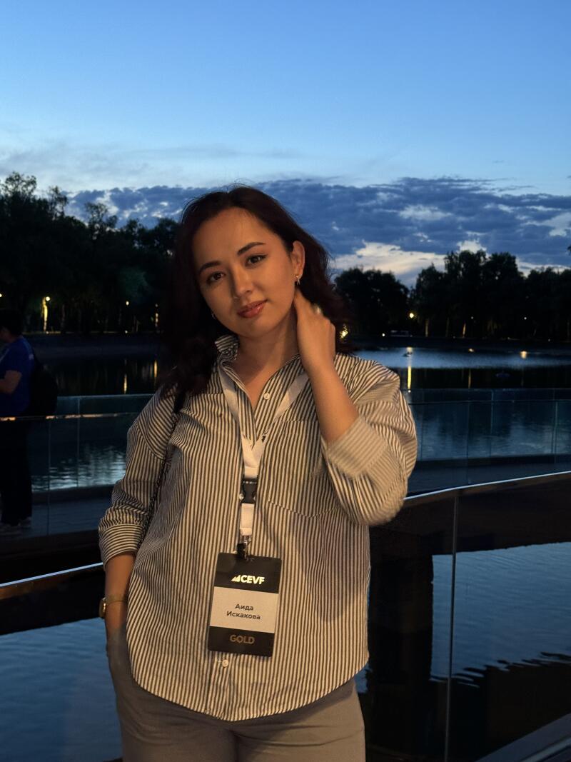 A woman wearing a striped shirt and a "GOLD" name tag that reads "ACEVF Аида Искакова" stands in front of a lake with trees and lights in the background under a twilight sky.