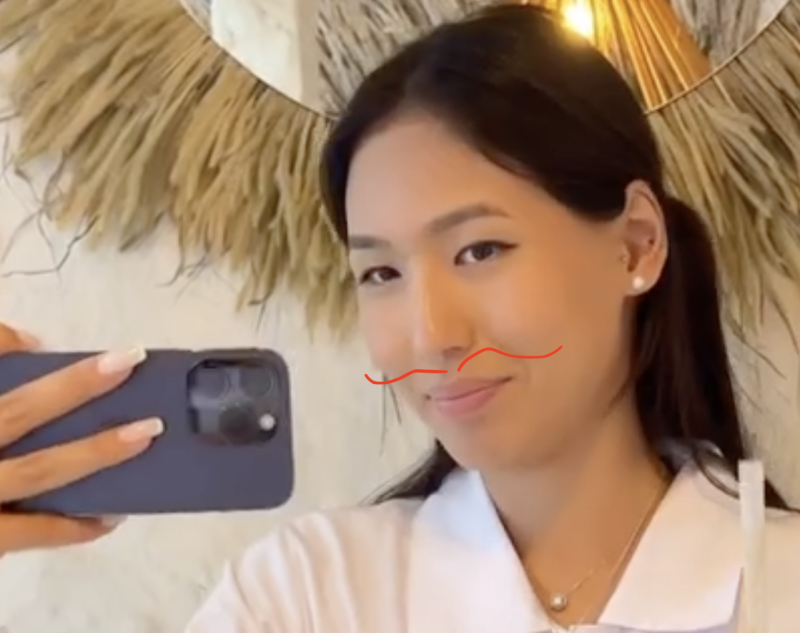 A woman with dark hair in a ponytail is taking a selfie with a dark-colored smartphone, wearing a white top and a delicate necklace.