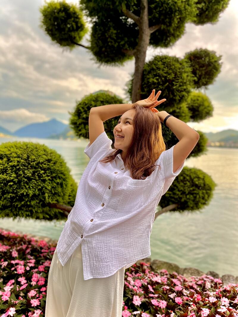 A woman in a white shirt and pants poses with her hands above her head in front of a flowerbed and a tree with round foliage, with a body of water and mountains in the background.