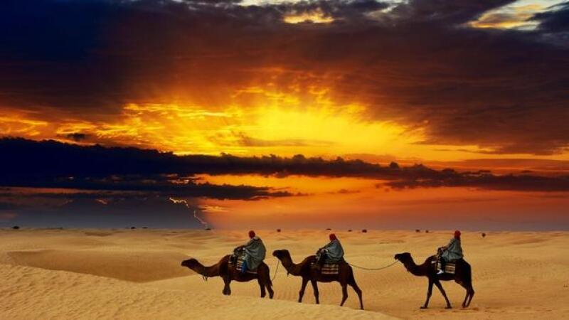 Three people are riding camels across a sandy desert at sunset, with a dramatic sky overhead.