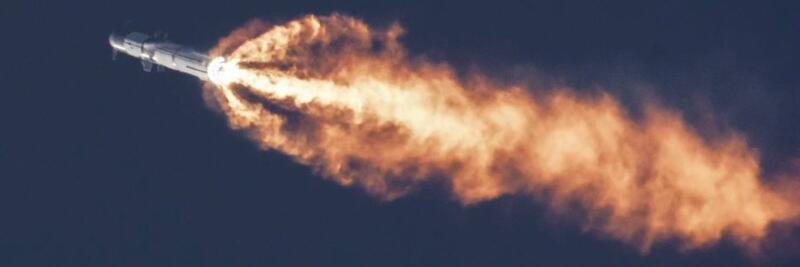A rocket is seen in flight, emitting a long plume of orange and yellow flames against a dark sky.